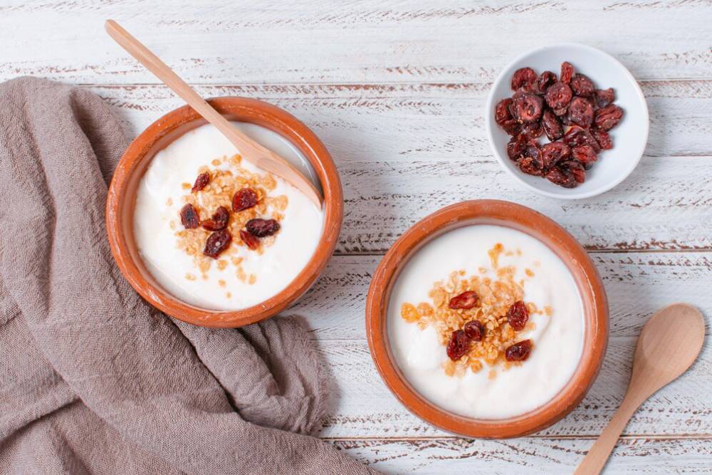 Two bowls of creamy yogurt topped with granola and dried fruits, accompanied by a small dish of more dried fruits and a wooden spoon.
