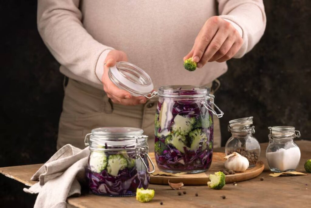 A person placing a piece of broccoli into a jar of pickled purple cabbage on a wooden table, with garlic and spices nearby.