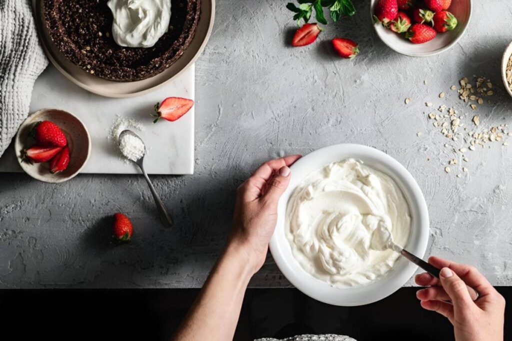 A person stirs yogurt in a bowl, surrounded by fresh strawberries, a pie crust, and a dish of coconut flakes.