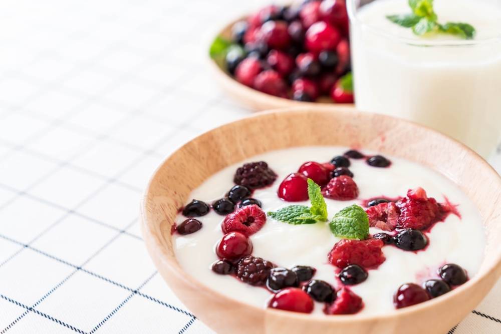 A wooden bowl filled with yogurt topped with fresh berries and a mint leaf, alongside a glass of yogurt and another bowl of mixed berries.