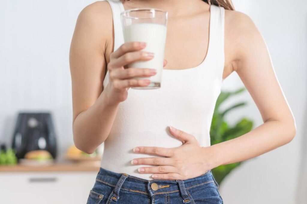 A woman in a white tank top holds a glass of milk while placing a hand on her stomach.