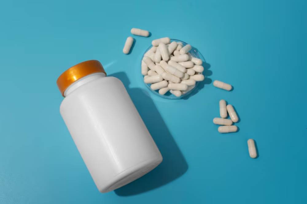 A white pill bottle with a gold lid is placed on a blue surface, surrounded by scattered and grouped white capsules in a dish.