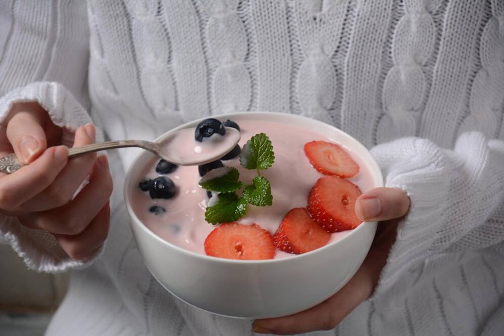 A person in a cozy white sweater holds a bowl of yogurt topped with strawberries, blueberries, and mint, ready to eat.