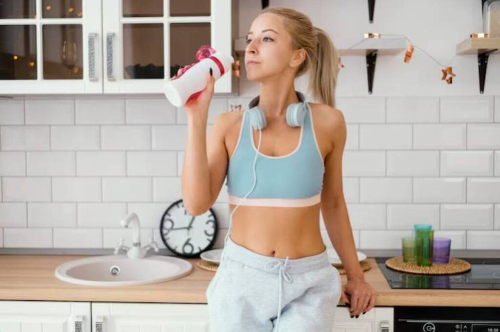 A fit individual in a sports bra and sweatpants holds a water bottle in a modern kitchen, adorned with colorful candles and decor.