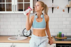 A fit individual in a sports bra and sweatpants holds a water bottle in a modern kitchen, adorned with colorful candles and decor.