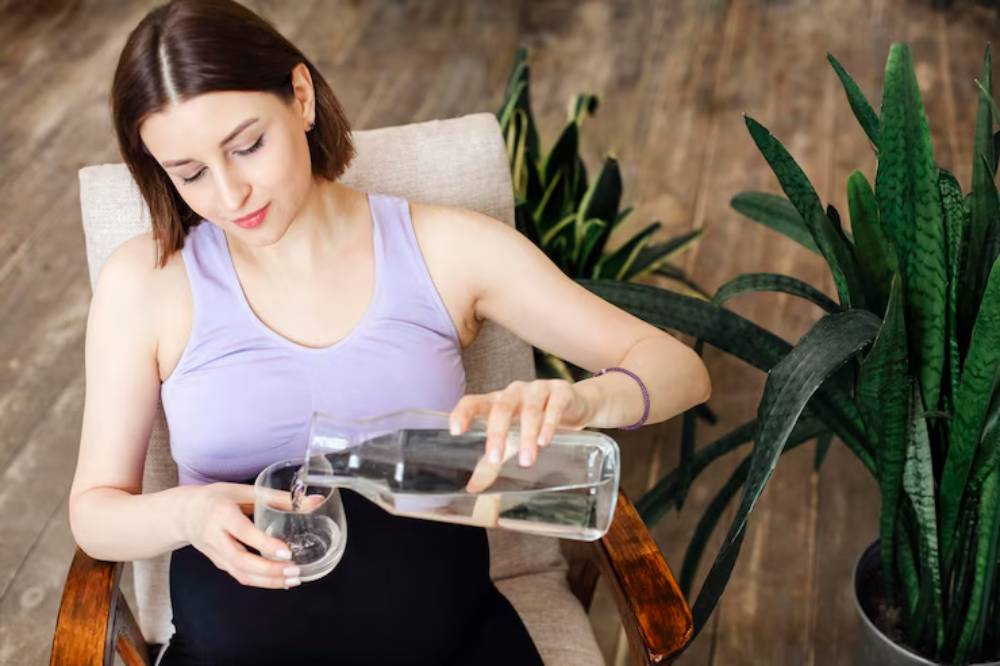 A person in a purple tank top pours water from a glass pitcher into a clear glass, surrounded by leafy plants in a cozy setting.
