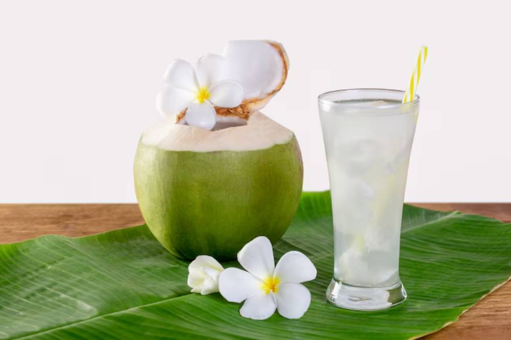 A fresh coconut with flowers beside it, next to a glass of coconut water with a straw, on a green banana leaf backdrop.
