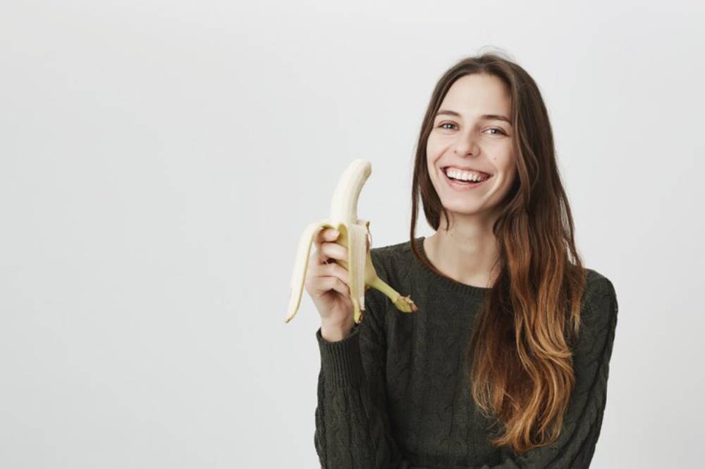 A person holds a peeled banana, wearing a dark sweater, against a plain white background.