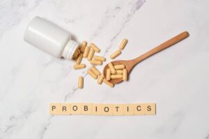 A wooden spoon holds yellow probiotic capsules next to a spilled white bottle and letter tiles spelling 'PROBIOTICS' on a countertop.