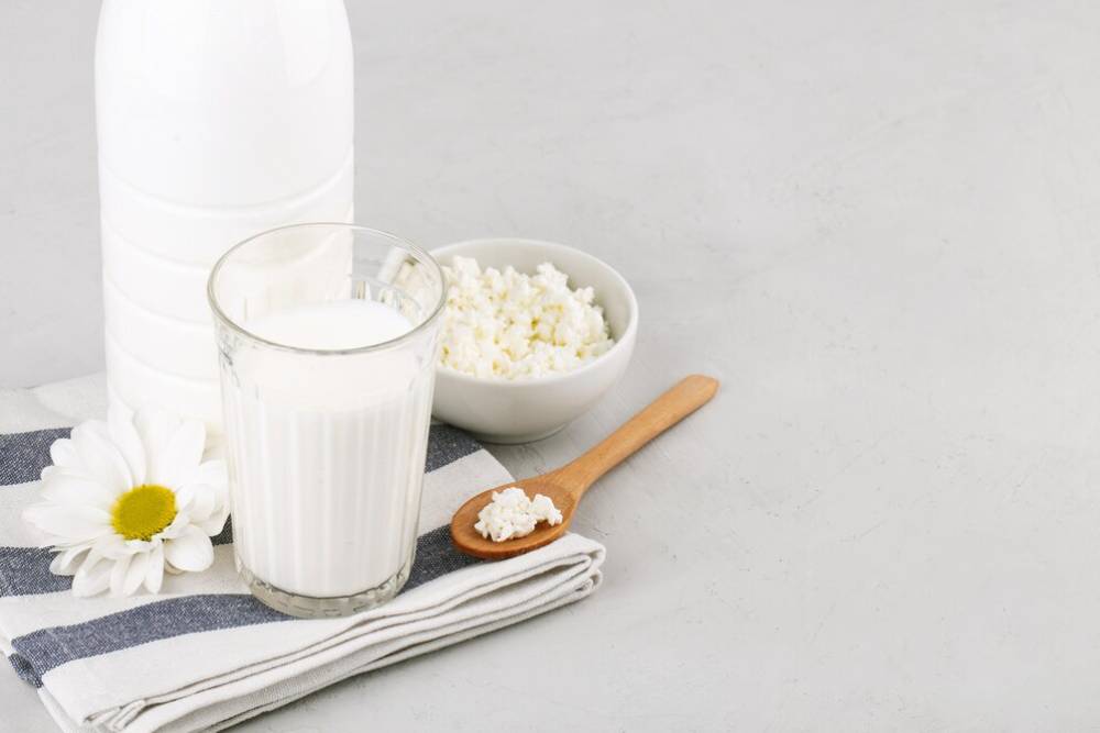 A glass of milk sits beside a bowl of cottage cheese, a wooden spoon, and a flower on a striped towel against a light background.