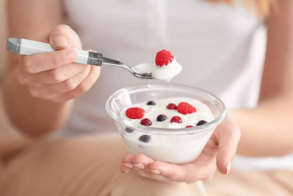 A person holds a bowl of yogurt topped with raspberries and blueberries, using a spoon to scoop a bite.