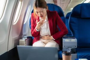 A woman in a red blazer sits on a plane, visibly uncomfortable, holding her stomach, while a laptop and coffee cup are nearby.