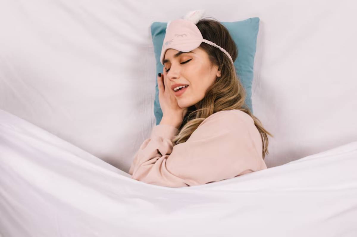 A person lies comfortably in bed, wearing a sleep mask and cozy loungewear, surrounded by soft pillows and blankets.