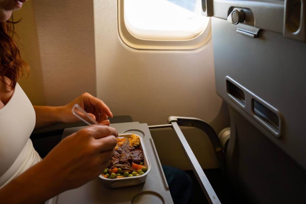 A passenger seated by an airplane window eats a meal from a tray, featuring meat and vegetables.