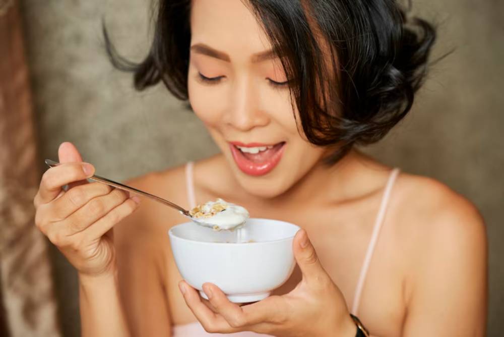 A person enjoys a bowl of yogurt topped with granola, holding a spoon, in a softly lit, neutral-toned setting.