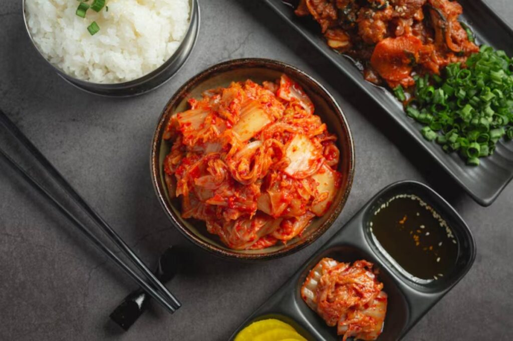 A bowl of spicy kimchi alongside white rice, sliced green onions, and a small dish with dipping sauce on a dark stone surface.