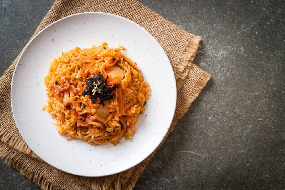 A plate of spicy kimchi fried rice topped with sesame seeds and seaweed, served on a textured burlap mat against a dark background.