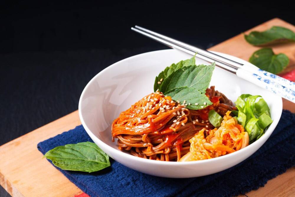 A white bowl filled with spicy noodle dish topped with sesame seeds and fresh herbs, alongside a small serving of kimchi and lettuce.