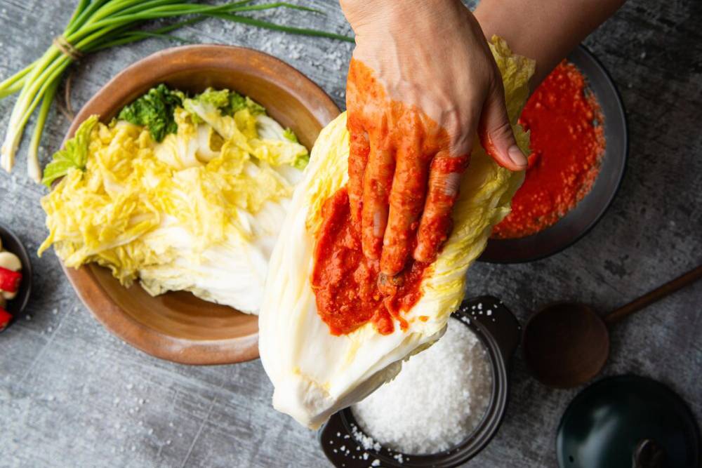 A hand applying red chili paste to a cabbage leaf, surrounded by fresh vegetables and bowls of seasoning on a textured surface.