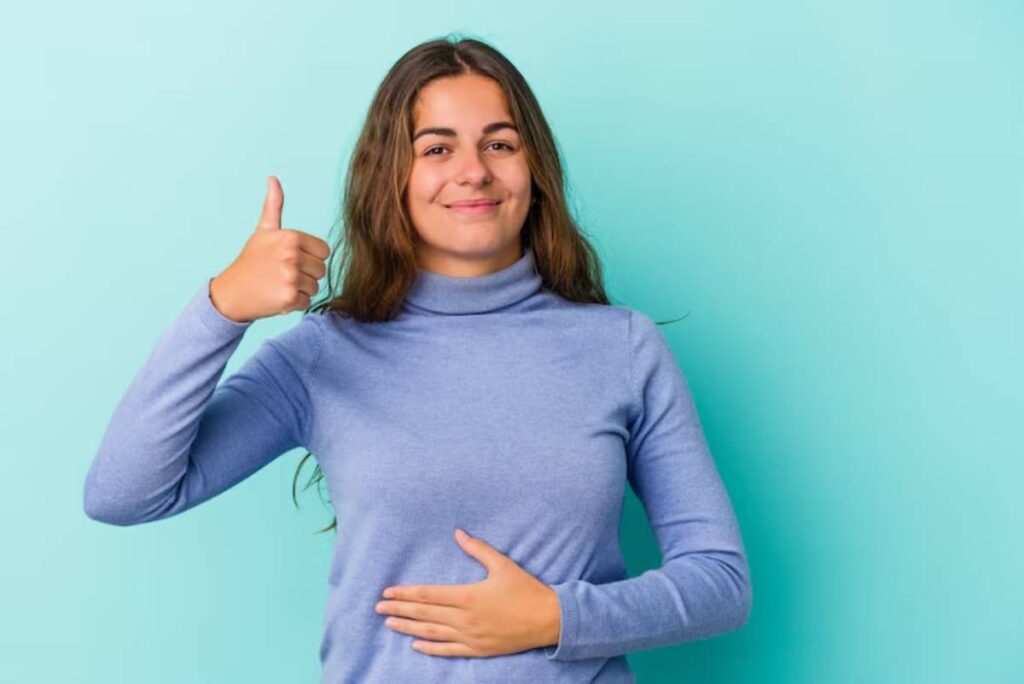 A woman in a blue turtleneck sweater gives a thumbs up while placing her hand on her stomach against a light turquoise background.