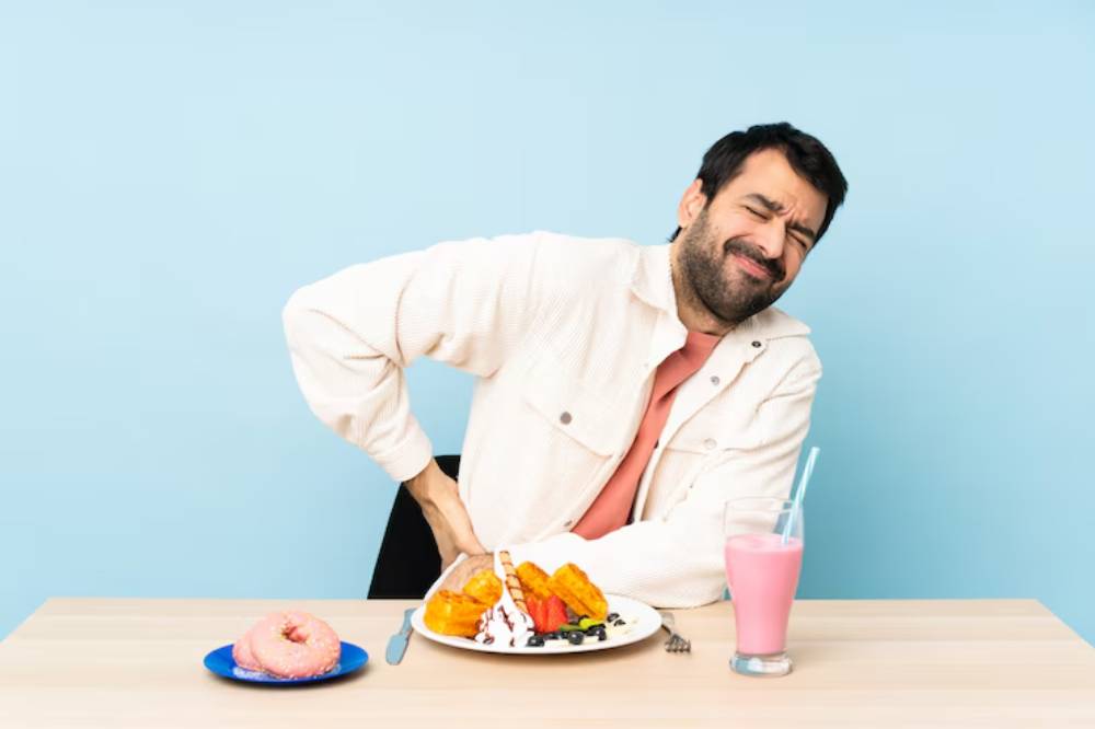 A person sits at a table with a plate of colorful pancakes, a pink milkshake, and a donut, against a light blue background.