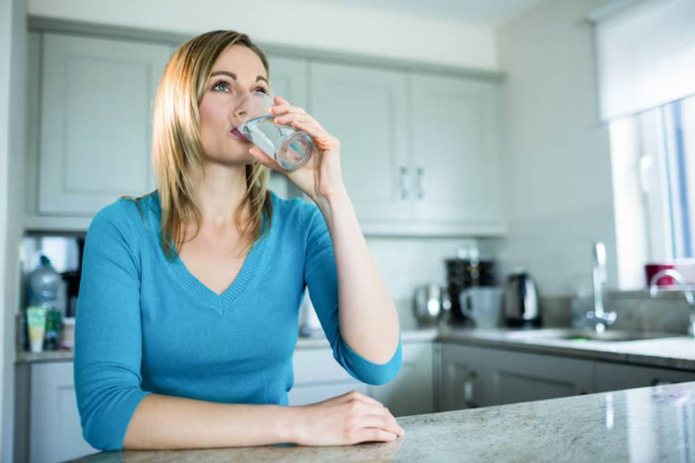 A woman in a blue sweater drinks water from a glass while seated at a kitchen countertop, surrounded by modern appliances.