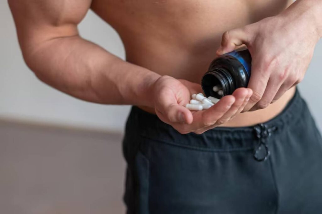 A muscular man in athletic shorts pours white supplements from a bottle into his palm, preparing for a workout.