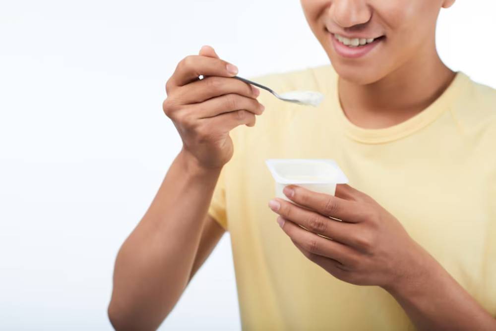 A smiling young man in a yellow shirt holds a spoonful of yogurt, preparing to eat from a small container.
