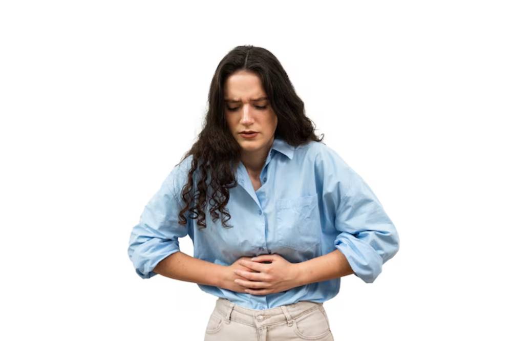 A woman in a light blue shirt stands with her hands clasped over her stomach, expressing discomfort against a plain white background.