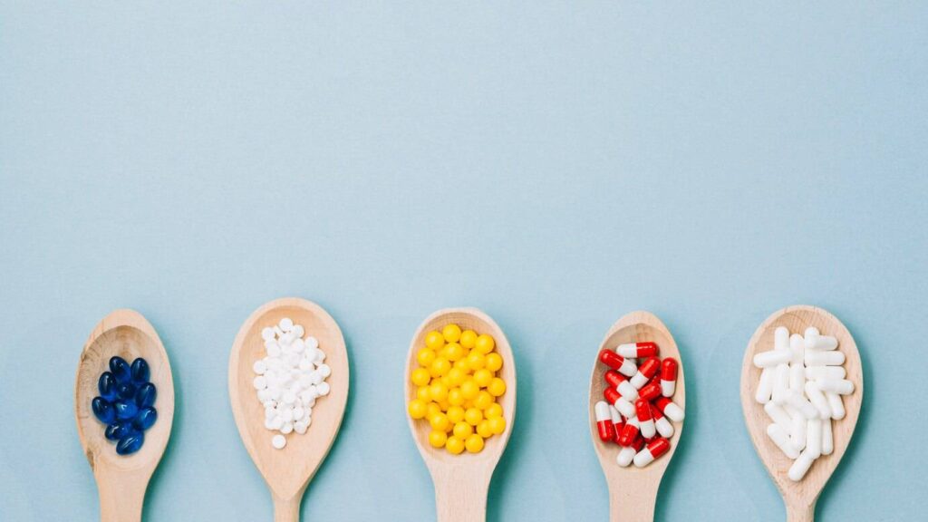 Five wooden spoons each filled with colorful pills: blue gel capsules, white tablets, yellow beads, red and white capsules, and more white tablets.