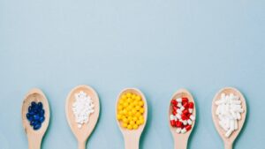 Five wooden spoons each filled with colorful pills: blue gel capsules, white tablets, yellow beads, red and white capsules, and more white tablets.