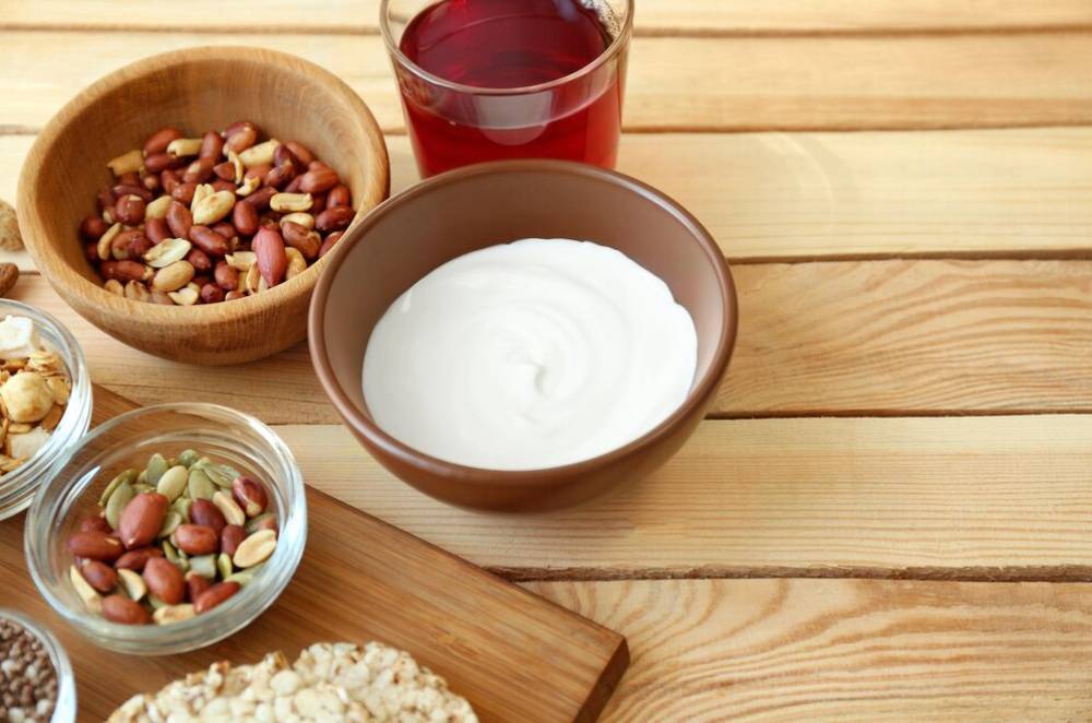 A bowl of yogurt beside mixed nuts, seeds, granola, and a glass of red drink, arranged on a wooden countertop.