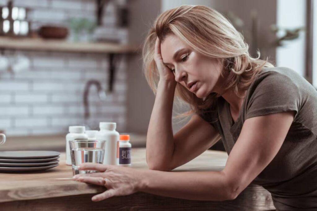A woman leans on a wooden table with a glass of water, surrounded by pill bottles and empty plates in a softly lit kitchen.