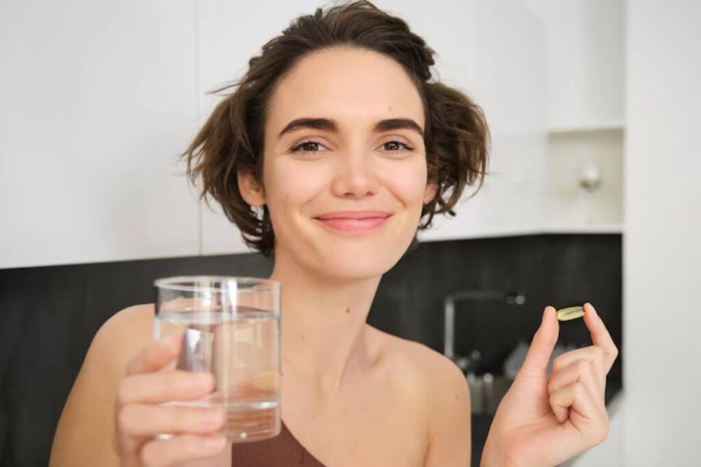 A person holds a glass of water in one hand and a supplement pill in the other, standing in a modern kitchen.