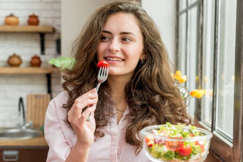 A woman with wavy hair and glowing skin holds a fork near a bowl of fresh salad, standing by a sunny window with plants and decorative jars in the background.