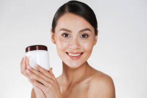 A woman holds a white jar with a brown lid, showcasing skincare against a plain light background.