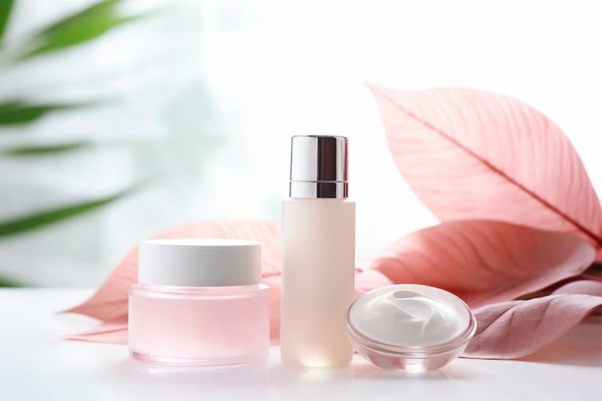 Three skincare products—a pink jar, a frosted bottle, and a white cream jar—set against delicate pink leaves and soft light.