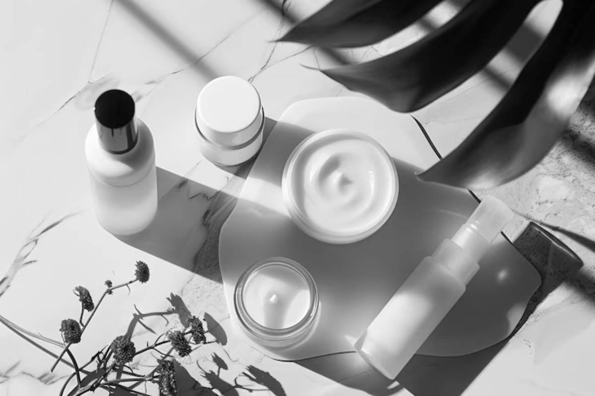 A flat lay of skincare products in black and white, featuring jars and bottles on a marble surface with soft shadows and a plant.