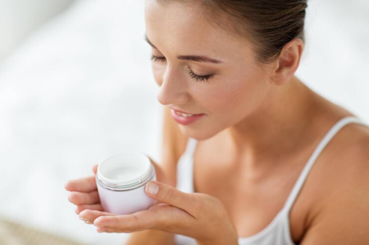 A woman in a white tank top holds a small jar of cream, contemplating its use in a softly lit indoor setting.