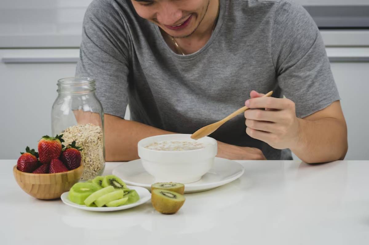 A man in a gray shirt smiles while stirring oatmeal in a bowl, with strawberries, kiwi, and oats displayed on the table.
