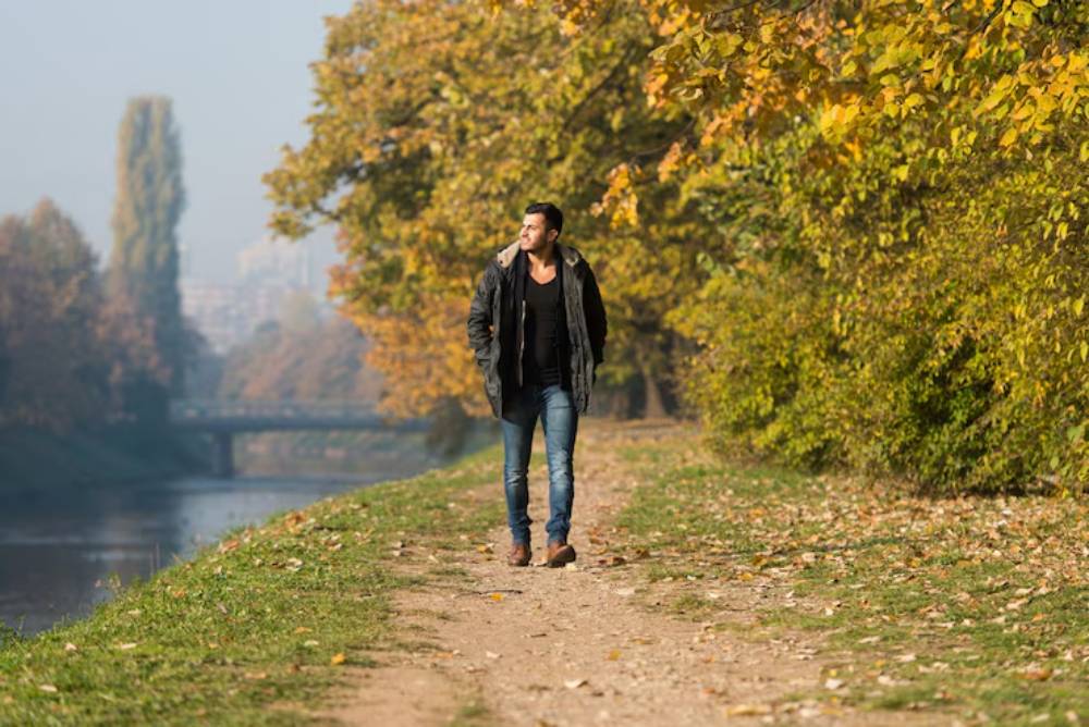 A person walks along a serene river path lined with autumn foliage, under a clear blue sky.