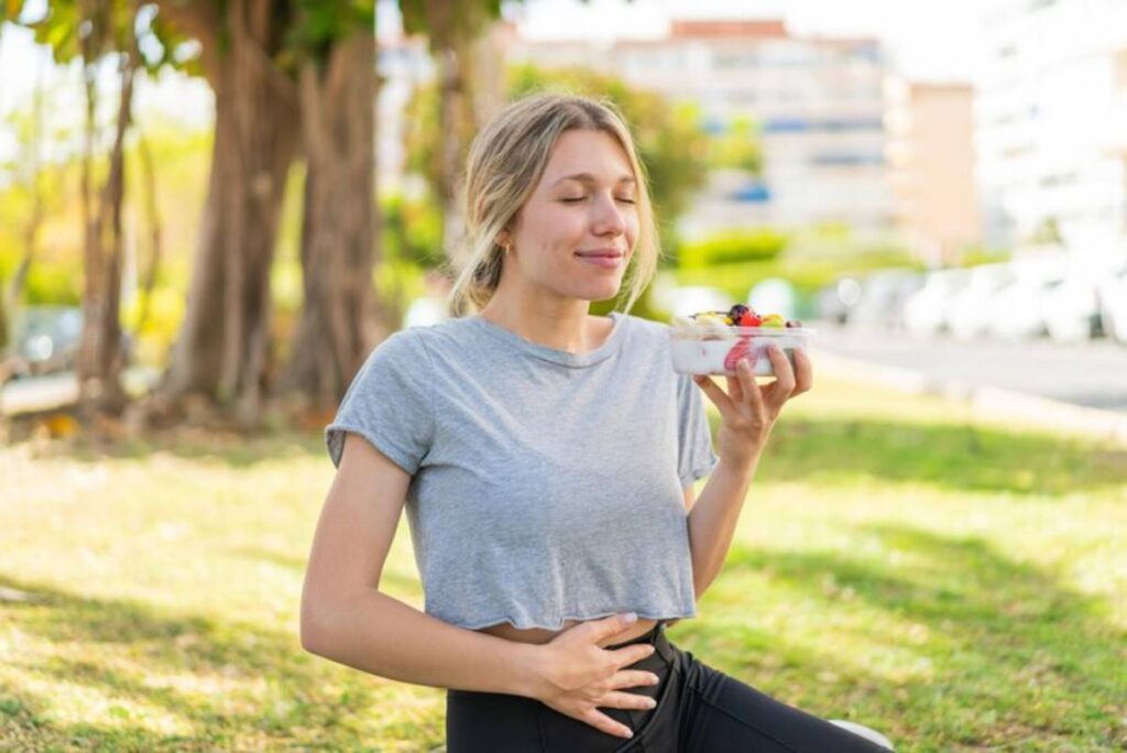 A woman in a gray crop top holds a bowl of fruit while sitting outdoors, with greenery and buildings in the background.