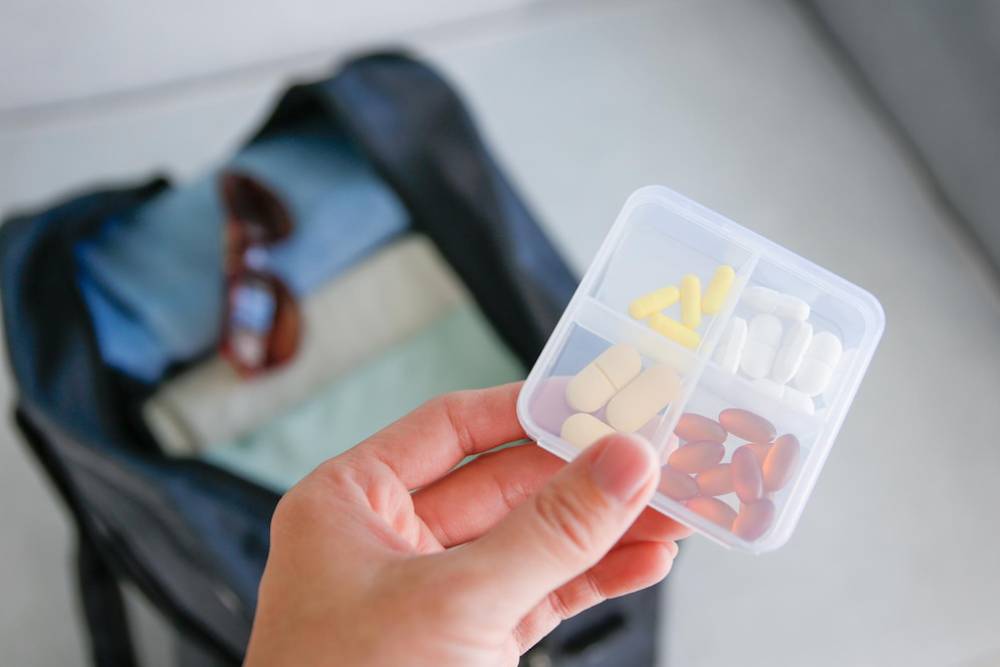 A hand holds a pill organizer with various colored capsules above an open bag containing clothes and sunglasses.