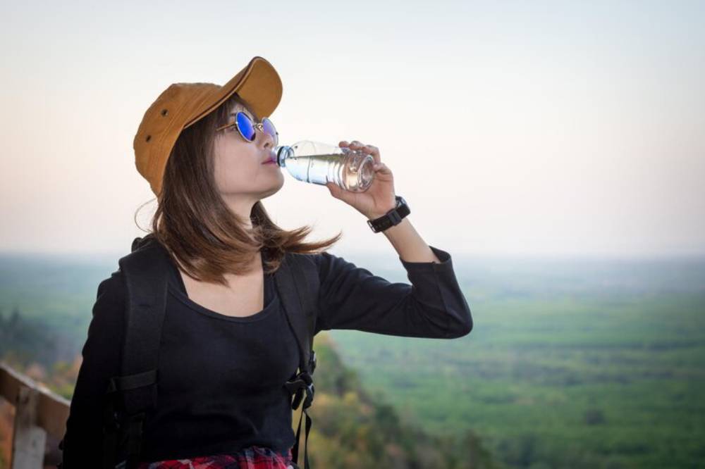 A person in a brown cap drinks water while enjoying a scenic view of lush greenery and distant hills during daylight.