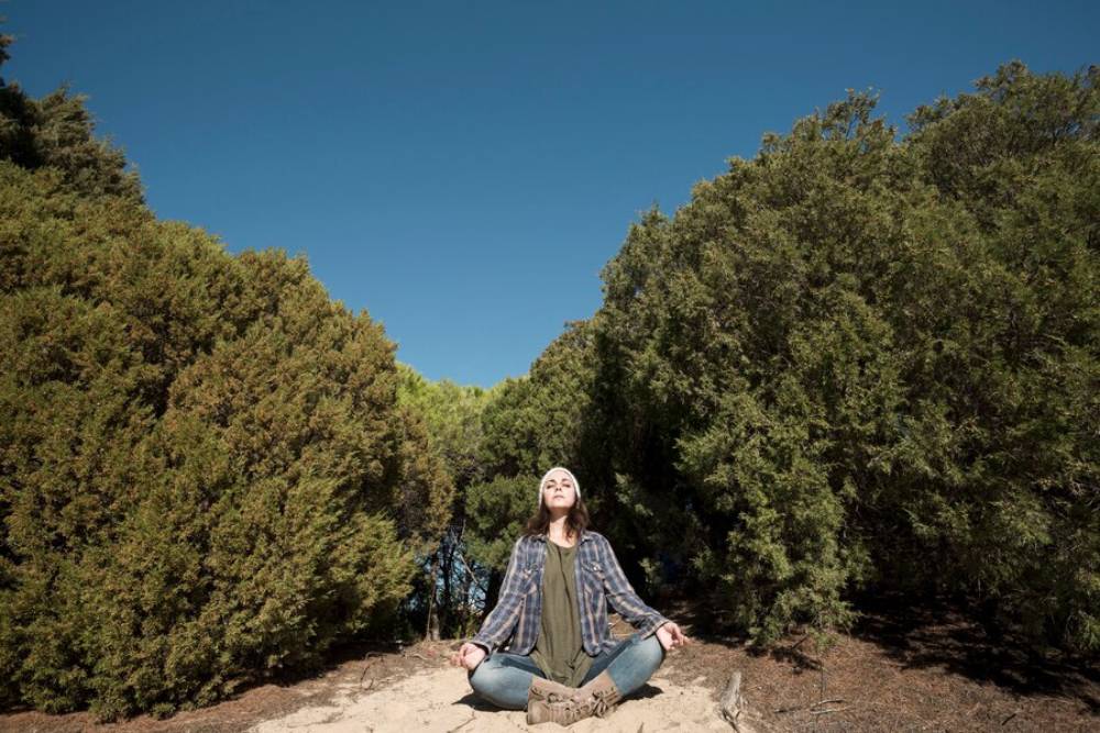 A person doing meditation on a sandy path surrounded by tall green shrubs under a clear blue sky, enjoying a moment of peace.