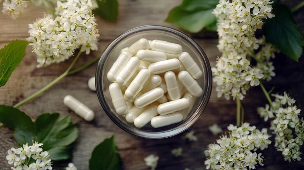 A glass bowl filled with white capsules surrounded by fresh white flowers and green leaves on a rustic wooden surface.