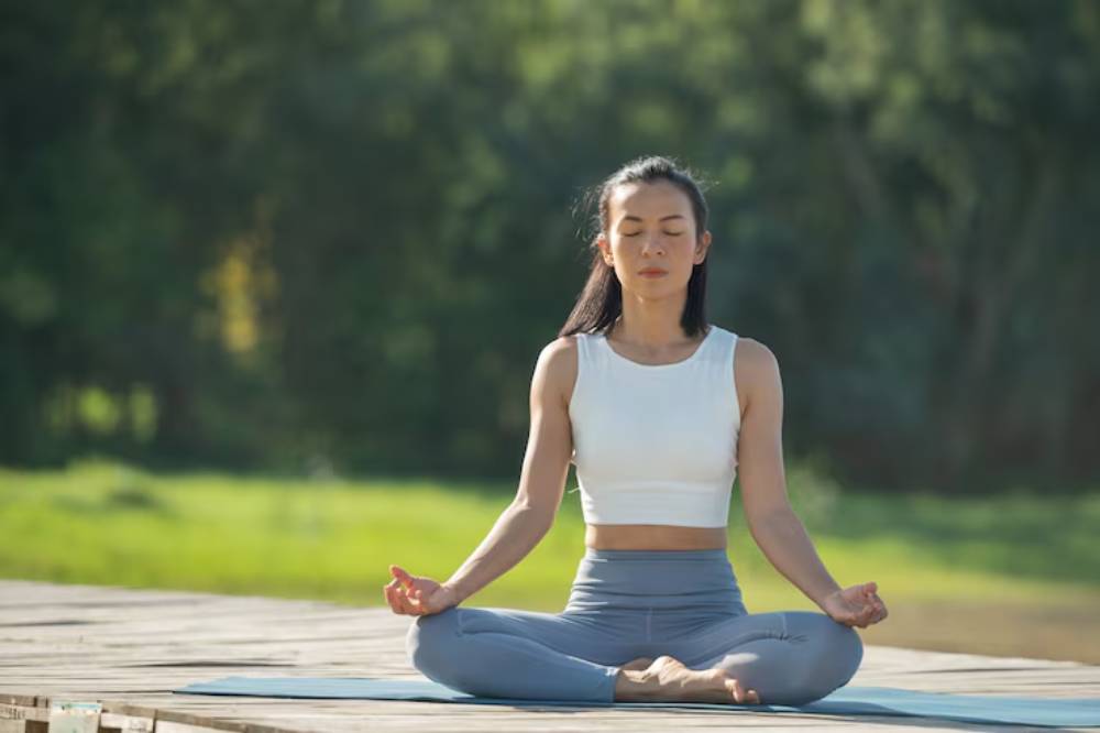 A person doing meditation in a serene outdoor setting, seated cross-legged on a mat, surrounded by lush greenery.